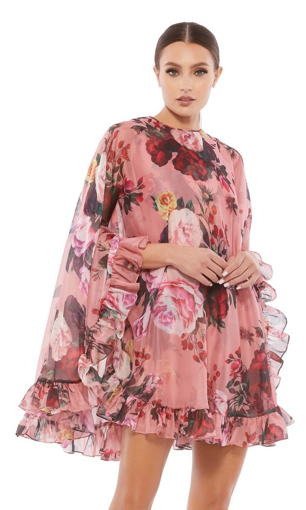 A woman wears the Mac Duggal 55404 Short Print Cape Hoco Dress, featuring a pink floral print, ruffled sleeves, and hem, standing against a white background.